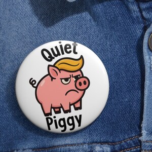 May include: A white button with a cartoon pig wearing a yellow toupee and a grumpy expression. The button reads "Quiet Piggy" in black text. The button is set against a blue denim background.