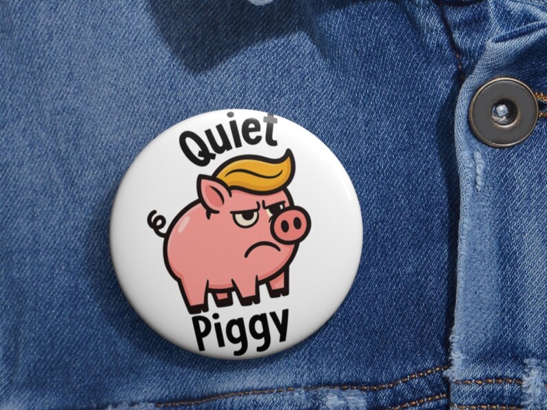 May include: A white button with a cartoon pig wearing a yellow toupee and a grumpy expression. The button reads "Quiet Piggy" in black text. The button is set against a blue denim background.
