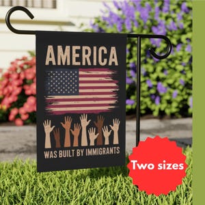 Peut inclure: Un drapeau de jardin noir avec un design de drapeau américain vieilli et le texte "America was built by immigrants". Le drapeau est affiché sur un support en métal noir devant une pelouse verte et des fleurs violettes.