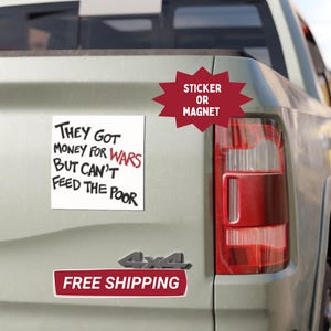 May include: A white square sticker or magnet with the text "THEY GOT MONEY FOR WARS BUT CAN'T FEED THE POOR" in black and red. The sticker is affixed to the back of a light-colored truck.