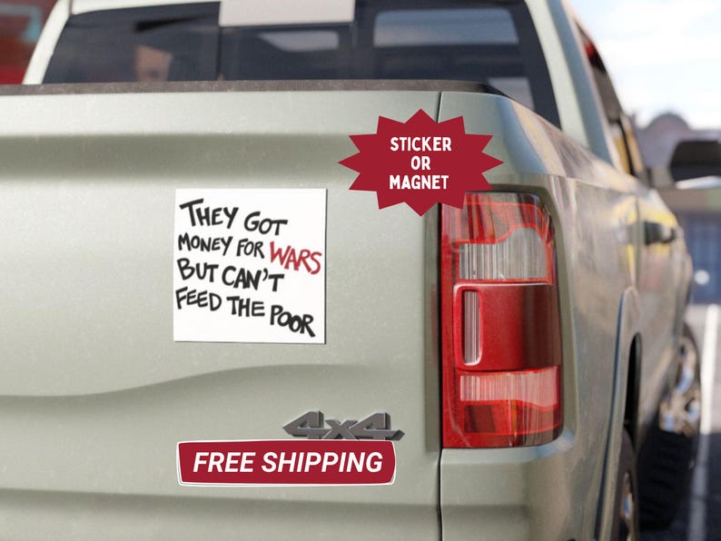 May include: A white square sticker or magnet with the text "THEY GOT MONEY FOR WARS BUT CAN'T FEED THE POOR" in black and red. The sticker is affixed to the back of a light-colored truck.