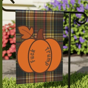 May include: A garden flag with a plaid background in shades of brown, green, and orange. An orange pumpkin graphic is centered on the flag, with the words "FUCK TRUMP" printed on the pumpkin. The flag is displayed on a black metal stand.