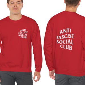 K&ouml;nnte beinhalten: Rotes Sweatshirt mit dem wei&szlig;en Text "ANTI FASCIST SOCIAL CLUB" auf der Vorder- und R&uuml;ckseite. Das Sweatshirt ist ein Rundhals-Stil und scheint aus einem weichen Material zu sein.