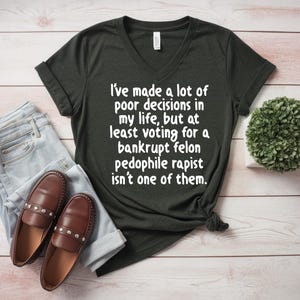 May include: Dark green V-neck t-shirt with white text: "I've made a lot of poor decisions in my life, but at least voting for a bankrupt felon pedophile rapist isn't one of them." Light blue jeans and brown loafers.
