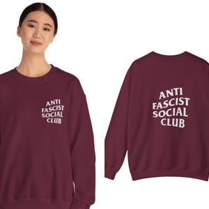 K&ouml;nnte beinhalten: Ein weinrotes Sweatshirt mit dem wei&szlig;en Text "ANTI FASCIST SOCIAL CLUB" auf der Vorder- und R&uuml;ckseite. Das Sweatshirt ist ein Rundhals-Modell und scheint aus einem weichen Material zu sein.