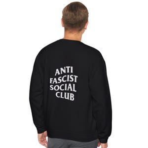 K&ouml;nnte beinhalten: Schwarzes Sweatshirt mit dem wei&szlig;en Text "ANTI FASCIST SOCIAL CLUB" auf dem R&uuml;cken. Das Sweatshirt ist einfarbig schwarz und scheint aus einem weichen Material zu sein.