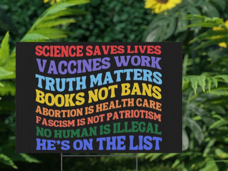 Op de afbeelding: Een zwart tuinbord met kleurrijke tekst. Het bord toont de zinnen: "Wetenschap redt levens, Vaccins werken, Waarheid doet ertoe, Geen boekenverboden, Abortus is gezondheidszorg, Fascisme is geen patriottisme, Geen mens is illegaal, Hij staat op de lijst."