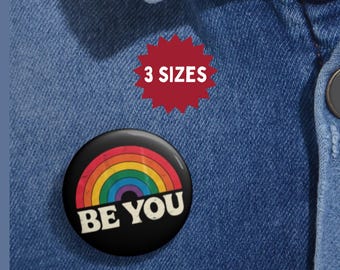 Può includere: Un bottone rotondo nero con un design arcobaleno e la scritta bianca "BE YOU". Sopra, una forma a stella rossa con il testo "3 SIZES". Il bottone è appuntato su una camicia di jeans blu.