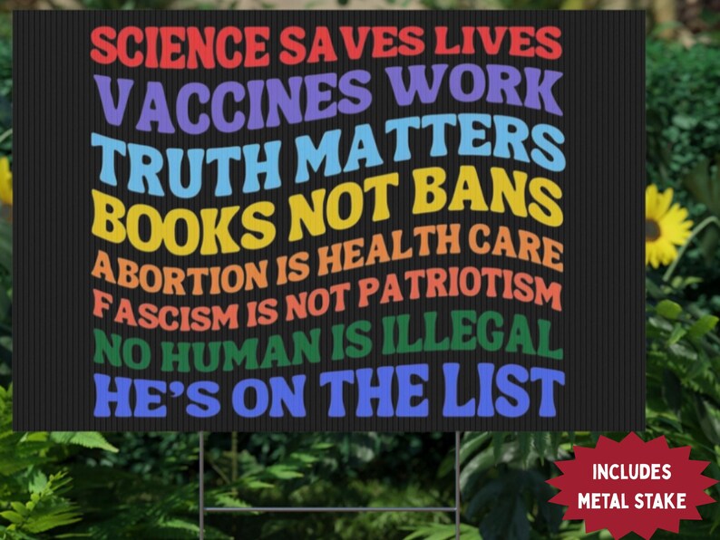 Op de afbeelding: Een zwart tuinbord met kleurrijke tekst met de tekst "WETENSCHAP REDT LEVENS, VACCINS WERKEN, WAARHEID DOET ERTOE, GEEN BOEKEN, ABORTUS IS GEZONDHEIDSZORG, FASCISME IS GEEN PATRIOTISME, GEEN MENS IS ILLEGAAL, HIJ STAAT OP DE LIJST." Inclusief een metalen paal.