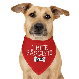 Peut inclure: Un chien beige porte un bandana rouge avec le texte "I BITE FASCISTS" en blanc. En dessous, un os blanc avec des empreintes de pattes noires. Le chien est centré sur un fond blanc.