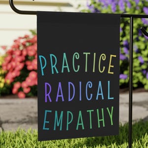 May include: A black garden flag with the words "PRACTICE RADICAL EMPATHY" in colorful, whimsical lettering. The flag is displayed outdoors, with a backdrop of green grass and vibrant flowers.