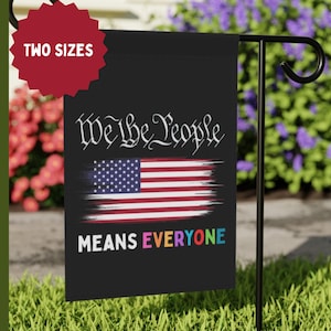 Op de afbeelding: Zwarte tuinvaandel met de tekst "We the People" en een ontwerp van de Amerikaanse vlag. De woorden "Means Everyone" zijn in regenboogkleuren. Een rood sterlabel leest "Two Sizes".