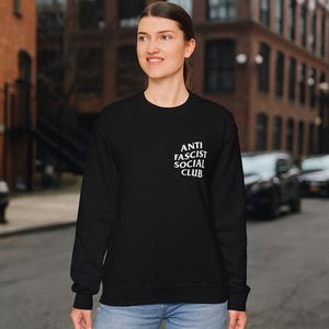 K&ouml;nnte beinhalten: Schwarzes Sweatshirt mit dem wei&szlig;en Text "ANTI FASCIST SOCIAL CLUB" auf der linken Brust. Das Sweatshirt wird von einer Person auf einer Stadtstra&szlig;e getragen.