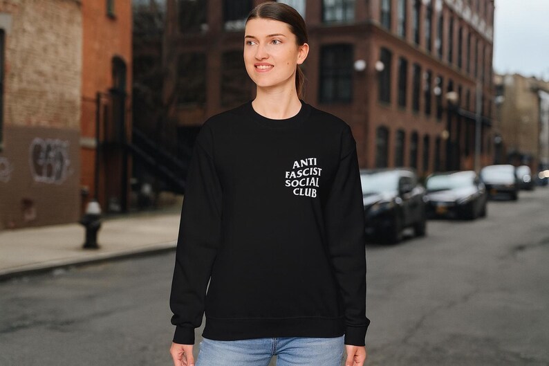 K&ouml;nnte beinhalten: Schwarzes Sweatshirt mit dem wei&szlig;en Text "ANTI FASCIST SOCIAL CLUB" auf der linken Brust. Das Sweatshirt wird von einer Person auf einer Stadtstra&szlig;e getragen.