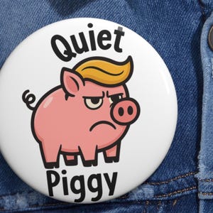 May include: A white button with a cartoon pig wearing a yellow toupee and an angry expression. The button reads "Quiet Piggy" in black text. The button is set against a denim background.