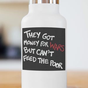 May include: White water bottle with a silver lid. The bottle has a black rectangle with white text that reads "They Got Money For Wars But Can't Feed The Poor". The word "Wars" is in red.