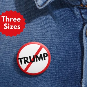 Puede incluir: Un botón rojo con texto blanco que dice "Three Sizes" y un botón rojo y blanco con una línea roja diagonal a través de la palabra "TRUMP" sobre un fondo de mezclilla azul.