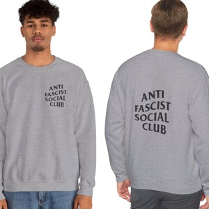 K&ouml;nnte beinhalten: Ein hellgraues Sweatshirt mit dem schwarzen Aufdruck "ANTI FASCIST SOCIAL CLUB" auf der Vorder- und R&uuml;ckseite. Das Sweatshirt hat einen Rundhalsausschnitt.