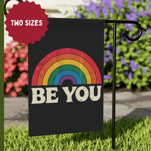 May include: Black garden flag featuring a rainbow design and the words "BE YOU" in white. The flag is displayed on a black metal stand in a garden setting. A red circular tag at the top left corner says "TWO SIZES".