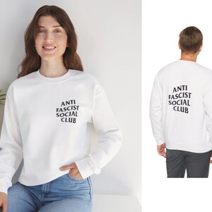 K&ouml;nnte beinhalten: Wei&szlig;es Sweatshirt mit dem Text "ANTI FASCIST SOCIAL CLUB" in Schwarz auf der Vorder- und R&uuml;ckseite. Das lang&auml;rmlige Sweatshirt hat einen Rundhalsausschnitt. Die Person auf dem Bild tr&auml;gt eine blaue Jeans.