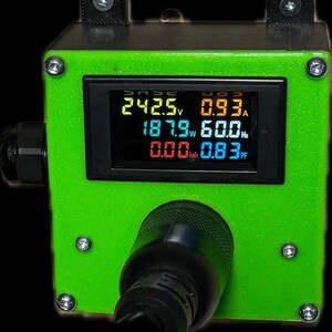 Może przedstawiać: Zielone urządzenie elektryczne z cyfrowym wyświetlaczem pokazującym odczyty napięcia, prądu, mocy, częstotliwości i współczynnika mocy. Urządzenie posiada czarną wtyczkę i jest zabezpieczone śrubami.