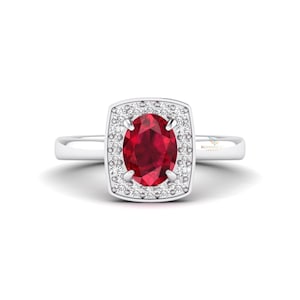 May include: A silver ring featuring a large, oval-cut, deep red gemstone surrounded by a square halo of smaller, clear gemstones. The ring's band is a simple, polished silver. The ring is set against a white background.