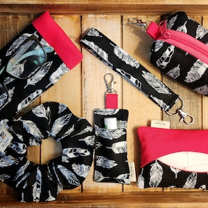 May include: A collection of black and white feather-patterned accessories with red accents, including a glasses case, scrunchie, wristlet, lip balm holder, zippered pouch, and tissue holder. The items are displayed on a wooden tray.