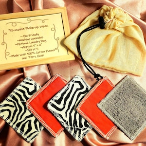 Puede incluir: Un juego de toallitas desmaquillantes reutilizables con una bolsa con cordón y un letrero de madera. Las toallitas presentan estampado de cebra, rojo y tela de rizo gris. El letrero dice "Re-usable Make-up Wipes". Cada toallita mide aproximadamente 10 cm x 10 cm.