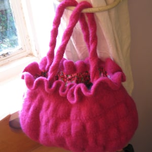 May include: A bright pink knitted bag with a ruffled top and two long handles. The bag is hanging from a white curtain.