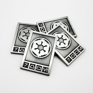 Puede incluir: Una colección de cinco fichas rectangulares de metal plateado. Cada ficha presenta un emblema circular en relieve con un diseño de estrella y el número "700M" en una fuente estilizada. Las fichas están dispuestas sobre un fondo blanco.