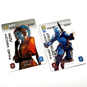 May include: Two rectangular game cards featuring character illustrations. The left card shows a blue-skinned figure labeled SPY TOKEN UNIT with RAID 2. The right card shows a figure in blue armor labeled MANDALORIAN TOKEN UNIT with SHIELDED.