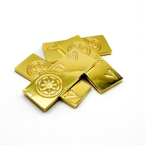 Puede incluir: Una colección de barras rectangulares doradas con diseños en relieve. Las barras presentan varios símbolos y formas, incluyendo un emblema circular y un '7' estilizado. Los objetos están dispuestos sobre una superficie blanca.