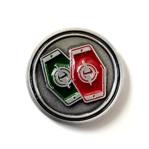 Puede incluir: Un pin redondo, de color plateado, con un borde en relieve. En el interior, se muestran dos emblemas en forma de escudo: uno verde y otro rojo, cada uno con un diseño plateado. El pin está sobre un fondo blanco.