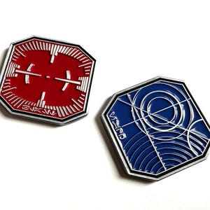 Puede incluir: Dos pines metálicos octagonales. Uno es rojo con líneas blancas y un diseño de cruz. El otro es azul con diseños circulares y de líneas blancas. Ambos tienen bordes plateados y texto.