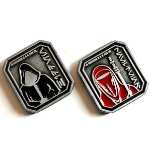 Puede incluir: Dos pines rectangulares plateados con un borde en relieve. Un pin presenta una figura negra, el otro una figura roja. Ambos pines tienen un diseño y texto en idioma extranjero.