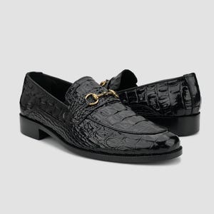 May include: Pair of black leather loafers with a crocodile texture. The shoes have a gold-colored metal buckle detail and a low heel. The glossy finish adds a touch of elegance to these classic slip-on shoes.