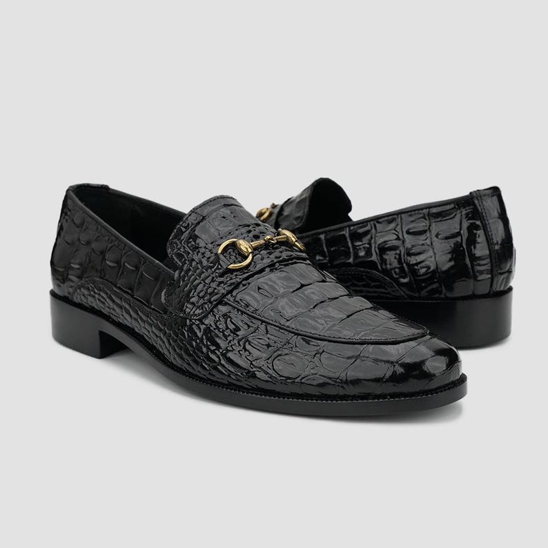 May include: Pair of black leather loafers with a crocodile texture. The shoes have a gold-colored metal buckle detail and a low heel. The glossy finish adds a touch of elegance to these classic slip-on shoes.