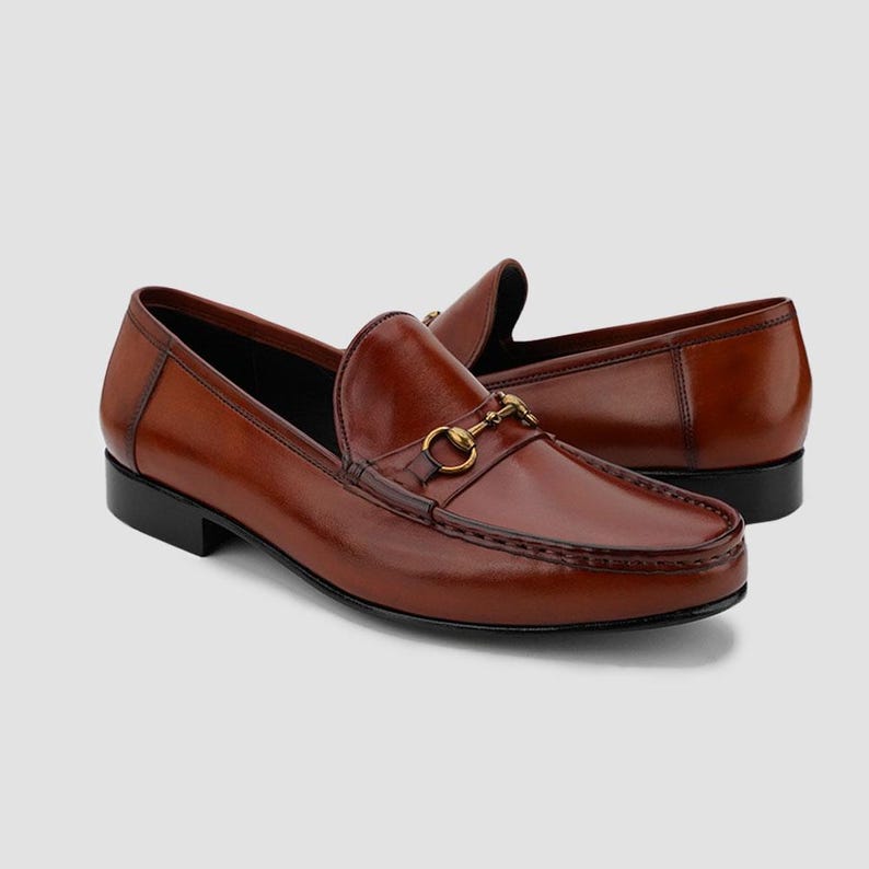 May include: A pair of brown leather loafers with a gold-colored metal horsebit detail. The shoes have a classic design with a rounded toe and a low heel. The leather has a polished finish and a dark brown color.