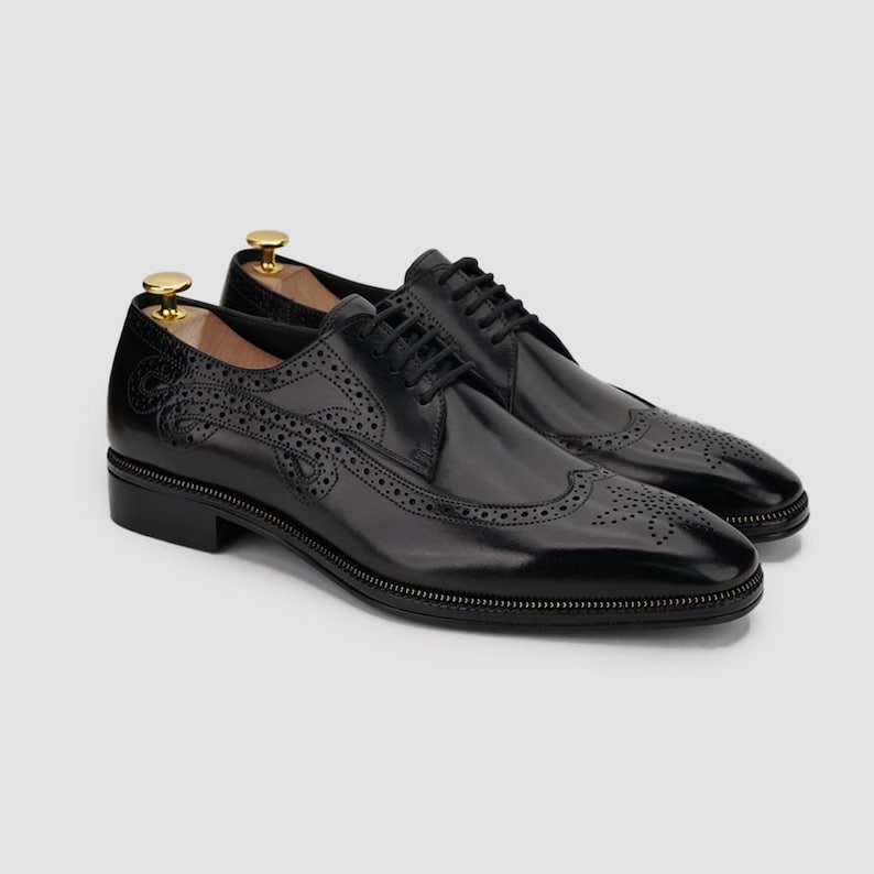 Puede incluir: Un par de zapatos de vestir de cuero negro con intrincados detalles de brogue y un acabado pulido. Los zapatos presentan un dise&ntilde;o cl&aacute;sico con cordones y se exhiben con hormas de madera. Las suelas tienen un sutil detalle de cremallera.