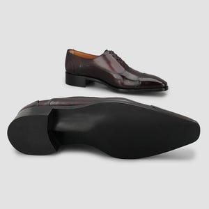 May include: A pair of dark burgundy leather oxford shoes with a cap toe and perforated detailing. One shoe is positioned upright showing the side profile and laces, while the other is placed in the foreground showing the smooth black sole.