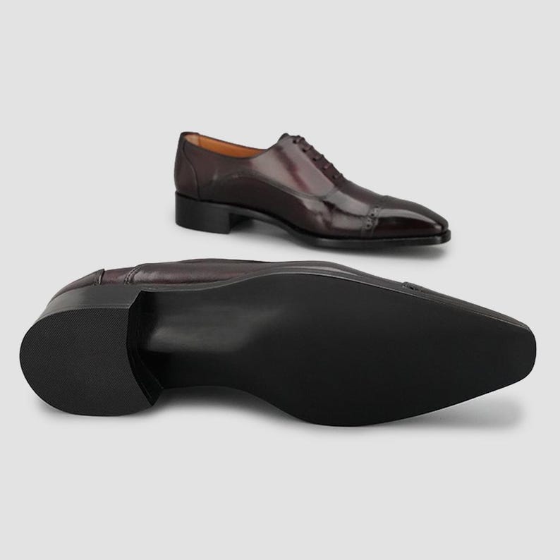 May include: A pair of dark burgundy leather oxford shoes with a cap toe and perforated detailing. One shoe is positioned upright showing the side profile and laces, while the other is placed in the foreground showing the smooth black sole.