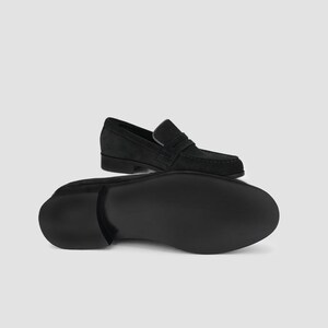 May include: Black suede loafers with a classic design. The image shows one shoe from the top and another from the bottom, showcasing the sole. The shoes have a slip-on style with a strap across the top.