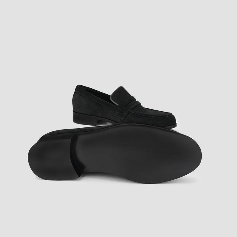 May include: Black suede loafers with a classic design. The image shows one shoe from the top and another from the bottom, showcasing the sole. The shoes have a slip-on style with a strap across the top.