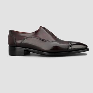 May include: A polished dark burgundy leather oxford shoe featuring a classic cap toe design with subtle brogue detailing along the seam, a low stacked heel, and a closed lacing system with thin matching laces on a smooth, sleek silhouette.