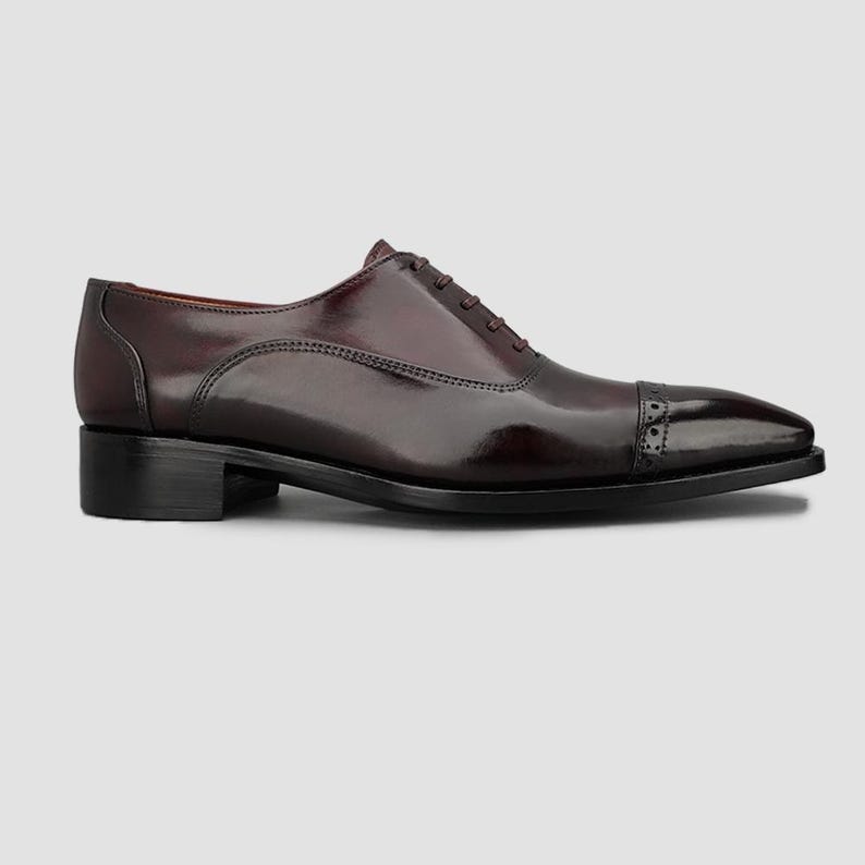 May include: A polished dark burgundy leather oxford shoe featuring a classic cap toe design with subtle brogue detailing along the seam, a low stacked heel, and a closed lacing system with thin matching laces on a smooth, sleek silhouette.