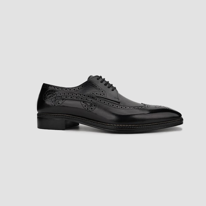 Puede incluir: Un zapato de vestir negro de cuero con dise&ntilde;o de ala. El zapato presenta perforaciones decorativas y una suela cosida. Los cordones son negros y el zapato tiene un tac&oacute;n bajo. El zapato est&aacute; sobre un fondo neutro.