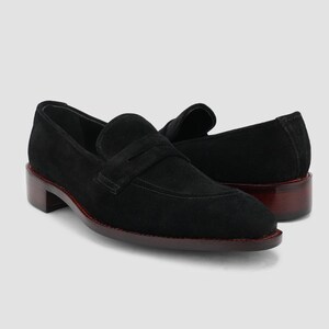 May include: A pair of black suede loafers with a classic penny strap design. The shoes have a low, stacked heel with a dark brown finish and a red trim around the sole. The loafers are displayed on a light gray background.