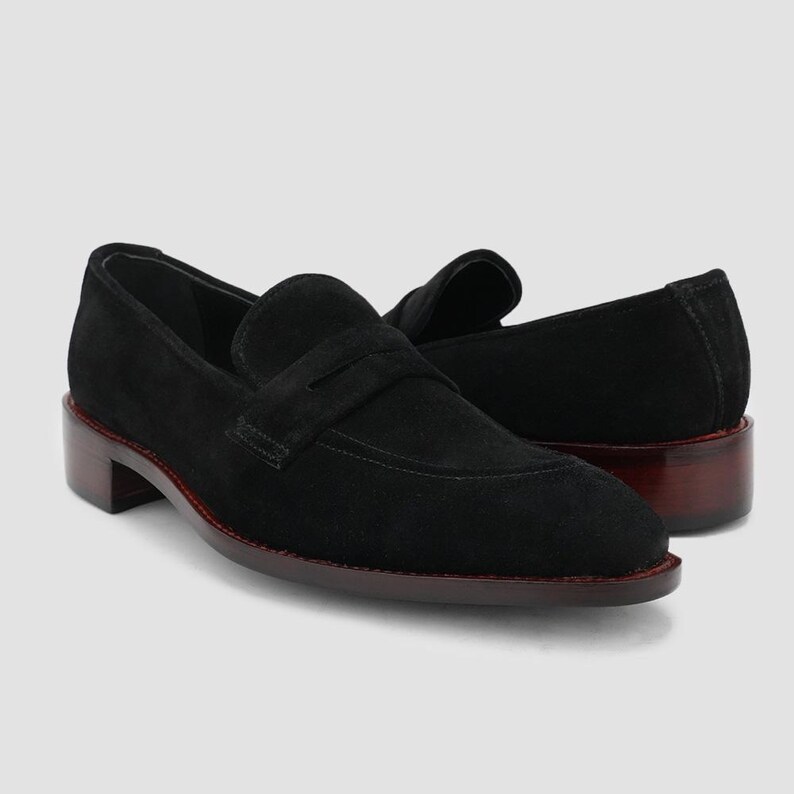May include: A pair of black suede loafers with a classic penny strap design. The shoes have a low, stacked heel with a dark brown finish and a red trim around the sole. The loafers are displayed on a light gray background.