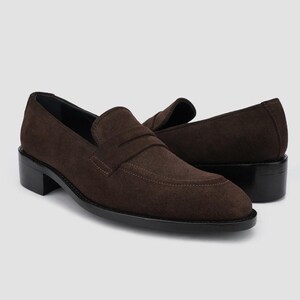 May include: A pair of brown suede loafers with a stacked heel. The shoes have a classic penny loafer design with a rounded toe and a stitched vamp. The soles and heels are black.