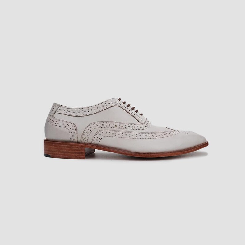 May include: A light gray leather oxford shoe with decorative perforations and brown laces. The shoe features a wingtip design and a stacked wooden heel. The shoe is a classic style, suitable for formal or business attire.